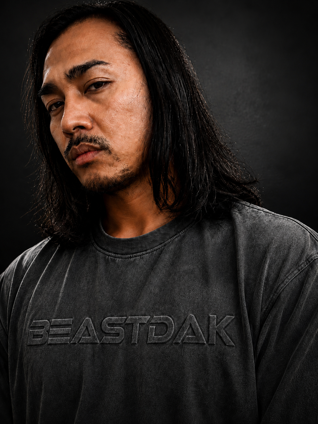 A man with long black hair and a beard wearing a dark gray crewneck t-shirt with the brand name 'BEASTDAK' embossed across the chest, set against a dark gray background.